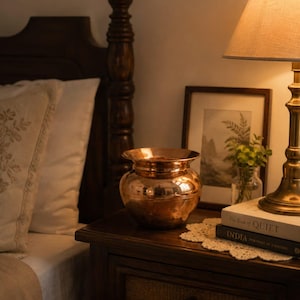 May include: A polished copper pot with a wide, flared rim and a rounded body. The metallic surface reflects the warm ambient light, showcasing a smooth, reflective finish. The vessel features a distinct neck and a curved base, standing as a standalone decorative object with a classic, rounded silhouette and a warm, reddish-brown metallic tone.