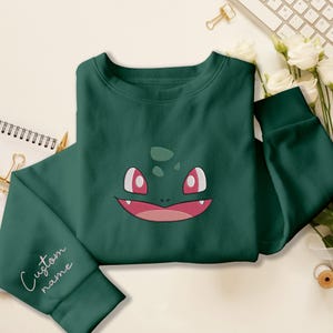 Puede incluir: Una sudadera de cuello redondo en color verde oscuro con un bordado que muestra la cara de una criatura con grandes ojos rosas y una boca ancha con dientes visibles. La manga izquierda incluye el texto bordado Custom name en cursiva.