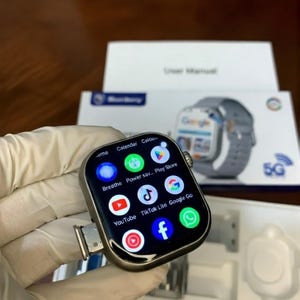 May include: A silver-toned smartwatch with a rectangular screen displaying various app icons including YouTube, TikTok Lite, Google Go, and Facebook. A metal SIM card tray is partially extended from the side of the watch casing, which is positioned in front of its original white product packaging.