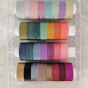 Può includere: Una collezione di sottili braccialetti metallici in un ampio spettro di colori, tra cui argento, oro, rosa, blu, verde e nero. I braccialetti sono organizzati in quattro file orizzontali su supporti cilindrici bianchi all'interno di un vassoio di plastica trasparente.