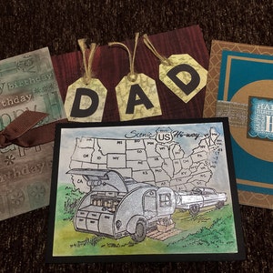 May include: A selection of handmade greeting cards. One card features a map of the United States with a vintage camper and car. Other cards have the words "Happy Birthday" and "DAD" on them. The cards are on a brown surface.