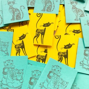 Könnte beinhalten: Eine Sammlung kleiner, quadratischer Karten in Türkis und Gelb mit Katzenillustrationen. Die türkisfarbenen Karten zeigen detaillierte Katzendesigns, während die gelben Karten einfachere Katzensilhouetten zeigen. Die Karten sind verstreut.