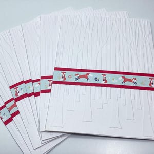 Może przedstawiać: Stos białych prostokątnych kart z wytłoczonym wzorem drzew leśnych. Każda karta jest owinięta poziomą jasnoniebieską opaską ozdobioną powtarzającym się wzorem pomarańczowych lisów, małych kwiatów i cienkich czerwonych pasków na krawędziach.