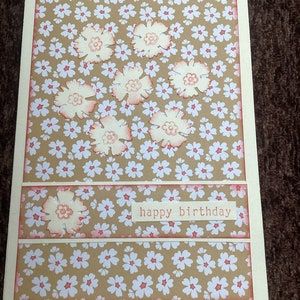 Op de afbeelding: Handgemaakte verjaardagskaart met een bloemenontwerp. De kaart heeft een beige achtergrond met een patroon van kleine witte bloemen. Grote, lichtroze bloemen zijn op de kaart geplaatst. De woorden "happy birthday" zijn gedrukt.