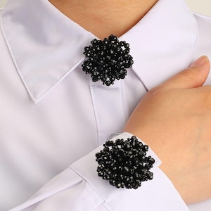 Puede incluir: Una camisa blanca de botones con dos racimos decorativos de cuentas negras. Un racimo está colocado en el cuello, mientras que el segundo está sujeto al puño de la manga. Las cuentas son oscuras, facetadas y de forma redonda.
