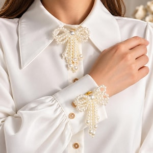Puede incluir: Una camisa blanca de manga larga con botones que presenta dos accesorios a juego en forma de lazo, elaborados con pequeñas perlas blancas artificiales. Un lazo de perlas está sujeto al cuello y el otro a la manga del puño.