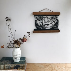 May include: A black and white paper cut-out art piece framed in a wooden frame. The art piece features two hands forming a fist with the word "Feminist" written on a banner.