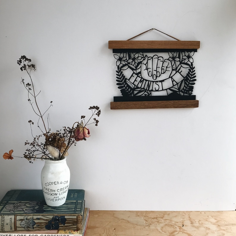 May include: A black and white paper cut-out art piece framed in a wooden frame. The art piece features two hands forming a fist with the word "Feminist" written on a banner.