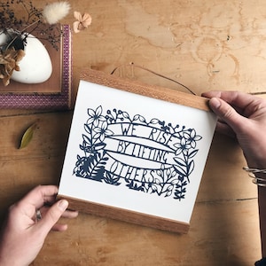 May include: A framed print with a black and white paper cut design of flowers and a banner with the text "We Rise By Lifting Others".