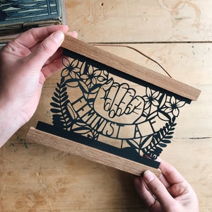 May include: A wooden frame with a black paper cutout design of two hands clasped together with the word "Feminist" written in a banner around them. The design is surrounded by floral and leaf motifs.