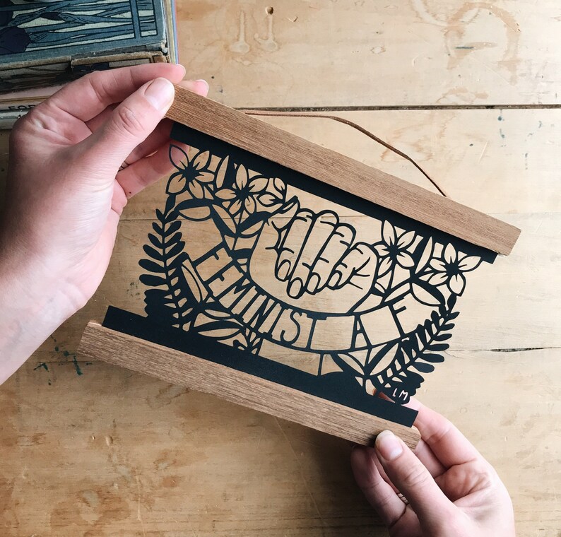 May include: A wooden frame with a black paper cutout design of two hands clasped together with the word "Feminist" written in a banner around them. The design is surrounded by floral and leaf motifs.