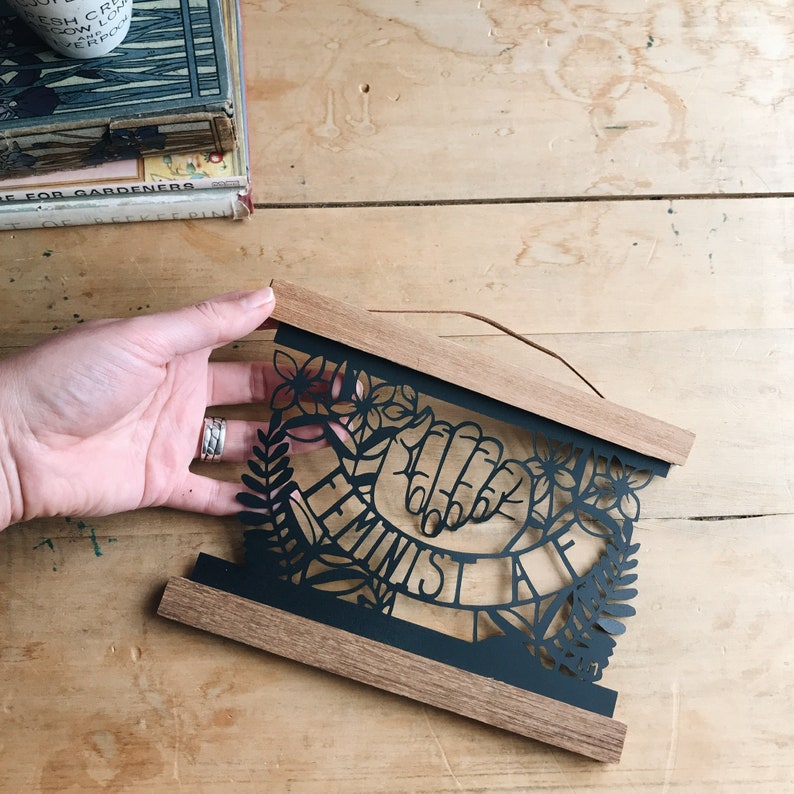 May include: Black and white paper cut-out art framed in wood. The art features two hands clasped together with the word "Feminist" written on a banner.
