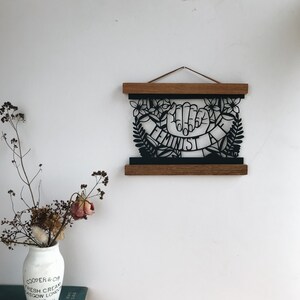 May include: Black and white paper cut-out art print of a hand with the word "Feminist" written on a banner. The print is framed in a wooden frame and hanging on a white wall.
