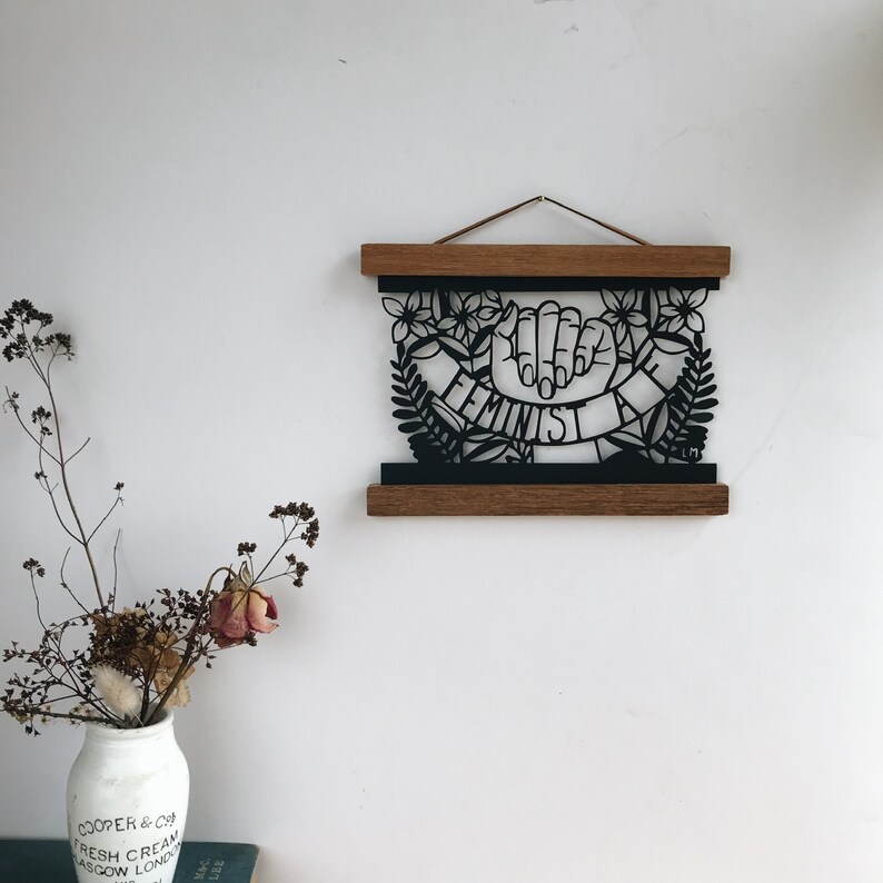 May include: Black and white paper cut-out art print of a hand with the word "Feminist" written on a banner. The print is framed in a wooden frame and hanging on a white wall.