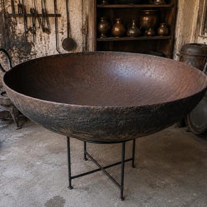 May include: A large, round, dark brown cast iron cauldron with a textured, weathered surface and two side handles. The heavy vessel is supported by a thin, black metal cross-legged stand, creating a contrast between the rustic bowl and the frame.