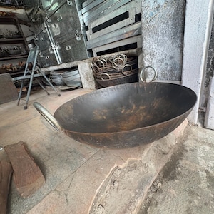May include: A large, round, dark-colored metal cooking pan with a wide, open basin and two curved metal handles attached to opposite sides. The surface of the pan shows a weathered, textured finish with visible signs of oxidation and dark patina.