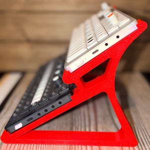 May include: A bright red, dual-tiered stand holding two electronic music synthesisers at an angled position. The top unit is white with orange knobs, while the bottom unit is black with white keys and various control buttons and ports.