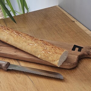 May include: A long, golden-brown baguette rests diagonally on a rectangular, rustic wooden serving board with a natural grain and a small black house-shaped icon. A serrated bread knife with a dark wooden handle lies beside the board.
