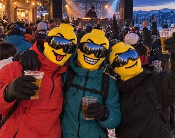 May include: Three individuals wearing bright yellow, molded head coverings resembling toy figures, paired with dark ski goggles. They are dressed in winter jackets and black gloves, each holding a clear plastic cup filled with a golden beverage.