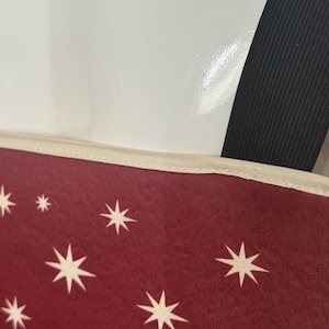 K&ouml;nnte beinhalten: Ein kastanienbraunes Stoffpaneel mit einem Muster aus wei&szlig;en achtstrahligen Sternen. Das Paneel hat eine cremefarbene Stoffkante an der Oberseite und ist an einem dicken schwarzen elastischen Band befestigt, das von der oberen rechten Ecke ausgeht.
