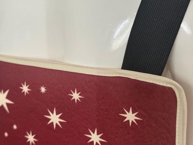 K&ouml;nnte beinhalten: Ein kastanienbraunes Stoffpaneel mit einem Muster aus wei&szlig;en achtstrahligen Sternen. Das Paneel hat eine cremefarbene Stoffkante an der Oberseite und ist an einem dicken schwarzen elastischen Band befestigt, das von der oberen rechten Ecke ausgeht.