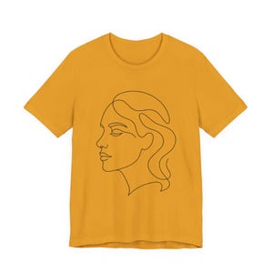 May include: A mustard yellow short-sleeved t-shirt featuring a black continuous line drawing of a human face in profile on the front. The design depicts the side view of a head with wavy hair, rendered in a single, fluid, unbroken black line.