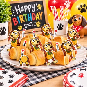 May include: A white plate filled with golden-brown crescent rolls, each topped with a decorative paper cutout of a cartoon dog character. The dog cutouts feature brown ears, a yellow body, and a small sign that says BARK! with a red heart.