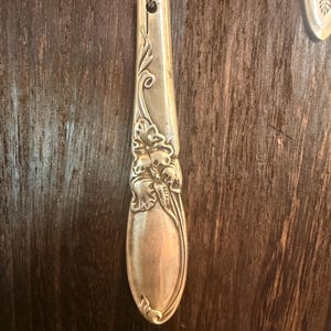 May include: A silver-toned pendant crafted from a repurposed spoon handle, featuring an embossed floral pattern along the stem. The pendant is suspended from a dark, braided cord, showcasing a metallic finish with intricate, raised botanical details.