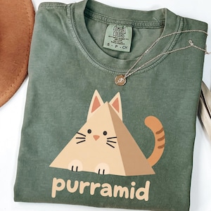 May include: A moss green crew neck t-shirt featuring a graphic of a tan triangular cat with a striped tail and the text purramid printed below. The shirt has a Comfort Colors label at the inner neckline and is paired with a thin gold chain necklace.