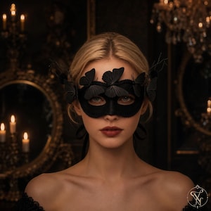 May include: A black masquerade mask featuring multiple three-dimensional butterfly embellishments across the top edge. The mask is crafted from a solid black material with a scalloped lace-like trim along the edges and includes thin black ribbons.