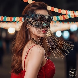 May include: A black lace masquerade mask featuring intricate embroidery, dark feathers, and decorative gold-toned accents. Long, delicate gold chains drape from the mask, creating a fringe effect that hangs down over the face and shoulders.