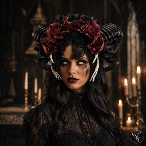 May include: A headpiece featuring large black curved horns, deep red fabric flowers, and white skeletal hand accents. The accessory is adorned with black lace detailing and is worn over long dark hair, creating a striking and detailed appearance.