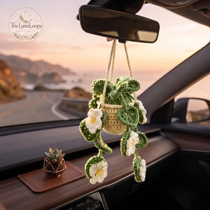 May include: A crocheted hanging plant ornament with a cream-colored pot and green leaves featuring white flowers with yellow centers, suspended by a cream cord from a car rearview mirror. A small succulent in a geometric copper holder sits nearby.