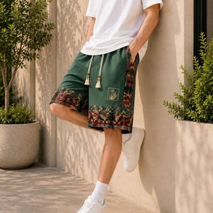 May include: Dark green shorts featuring a wide, multicolored geometric patterned hem and side panel. The shorts include a drawstring waist with cream-colored tassels and a small, square gold-outlined graphic print on the front left leg.