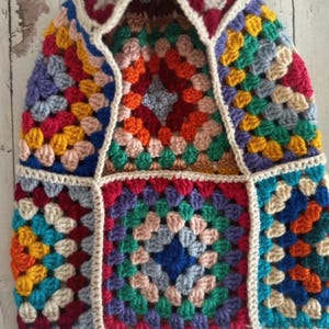 Peut inclure: Un bonnet en crochet composé de plusieurs carrés granny multicolores. Les carrés présentent des teintes vives de rouge, orange, jaune, vert, bleu et violet, le tout assemblé avec une bordure en fil de couleur crème sur les bords.