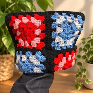 Könnte beinhalten: Ein Fischerhut aus gehäkelten Granny Squares in Schwarz, Rot, Rosa und Hellblau. Der Hut zeigt ein wiederkehrendes Muster aus diesen Quadraten, die mit schwarzem Garn verbunden sind, und bildet so ein strukturiertes, mehrfarbiges Accessoire.