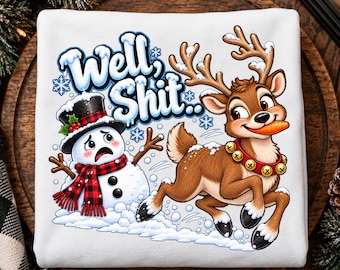 May include: A white sweatshirt featuring a graphic print of a cartoon snowman wearing a black top hat and red plaid scarf, and a brown reindeer with a red jingle bell collar holding a carrot in its mouth, with the text Well, Shit.. above them.