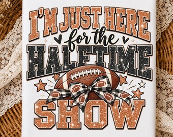 May include: A white t-shirt featuring the text I'M JUST HERE for the HALFTIME SHOW in copper glitter and black glitter lettering. The design includes a brown football with a black and white buffalo plaid bow and small copper stars.