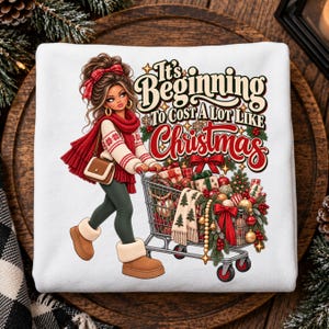 May include: A white sweatshirt featuring a graphic of a person with brown hair wearing a red scarf, white sweater, and tan boots, pushing a shopping cart filled with Christmas gifts, ornaments, and greenery, with the text It's Beginning To Cost A Lot Like Christmas.