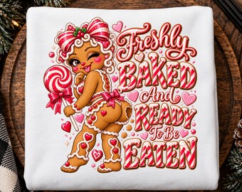 May include: A white sweatshirt featuring a graphic of a winking gingerbread character with a red and white striped bow, holding a lollipop. The text reads Freshly BAKED And READY To Be EATEN in red and white candy cane style lettering with hearts.