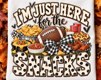 May include: A white t-shirt featuring a graphic print with the text I'M JUST HERE FOR THE SNACKS. The design includes a brown football with a black and white buffalo plaid bow, bowls of nachos, cheese dip, salsa, chicken wings, and red cups.