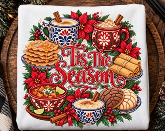 May include: A white sweatshirt featuring a colorful graphic with the text Tis The Season, surrounded by illustrations of mugs, cinnamon sticks, poinsettias, a stack of pancakes, tamales, a bowl of soup, and sweet bread rolls.