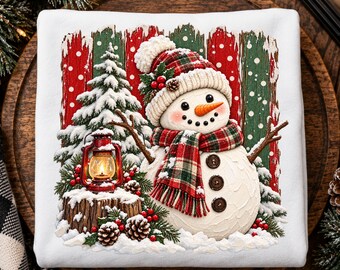 May include: A white sweatshirt featuring a graphic of a snowman wearing a red and green plaid scarf and matching hat. The snowman stands beside a snow-covered pine tree, a lit red lantern on a tree stump, and red berries and pinecones.