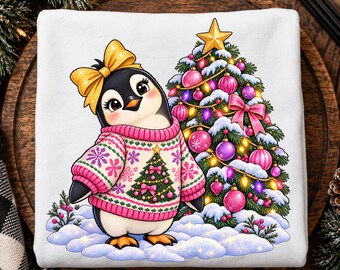 May include: A white sweatshirt featuring a graphic of a penguin wearing a pink and white snowflake-patterned sweater with a Christmas tree design and a yellow bow on its head, standing next to a decorated Christmas tree in the snow.