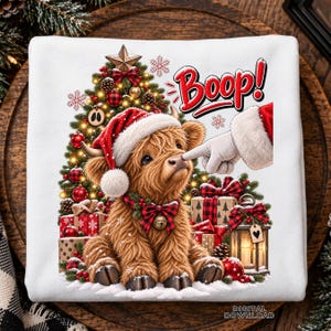 May include: A white sweatshirt featuring a graphic of a fluffy brown Highland cow wearing a Santa hat and a plaid bow with a bell. A gloved finger touches the cow's nose under the text Boop! A Christmas tree and gifts are in the background.