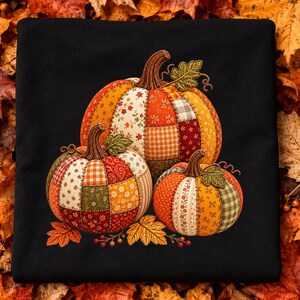 May include: A black t-shirt featuring a graphic of three pumpkins designed with a patchwork quilt pattern. The pumpkins are rendered in various shades of orange, red, green, and cream with floral and checkered prints, accented by brown stems and leaves.