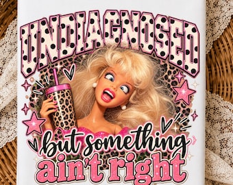 May include: A white t-shirt featuring a graphic of a blonde doll with a wide-eyed expression holding a leopard print tumbler. The text reads UNDIAGNOSED in black polka dots and but something ain't right in pink glitter. Pink stars and hearts surround the doll.