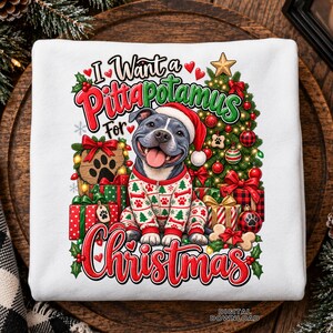 May include: A white sweatshirt featuring a graphic of a gray pit bull wearing a Santa hat and holiday pajamas. The dog sits before a decorated Christmas tree and presents. The text reads I Want a Pittapotamus For Christmas in red and green.