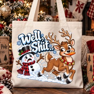 May include: A beige canvas tote bag featuring a graphic print of a snowman wearing a black top hat and red plaid scarf, and a reindeer with a bell collar holding a carrot in its mouth. The text "Well, Shit..." is printed in blue above them.