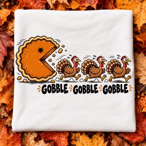 May include: A white t-shirt featuring a graphic of a large orange pumpkin pie shaped like a video game character chasing three brown turkeys. Below the graphic, the text GOBBLE GOBBLE GOBBLE is printed in a bold, black, rounded font.